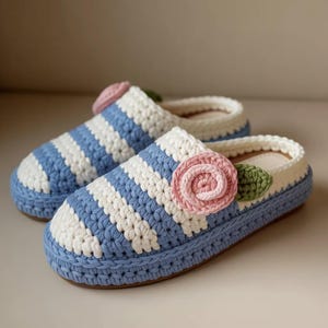 Puede incluir: Un par de zapatillas de ganchillo hechas a mano. Las zapatillas presentan rayas horizontales azules y blancas, una suela azul claro y una aplicación de rosa rosa con una hoja verde. El interior es de color beige claro.