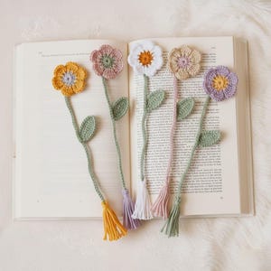 以下が含まれることがあります： 黄色、ピンク、白、紫など、さまざまな色の手作りのかぎ針編みの花のブックマークのコレクション。各ブックマークは、花の頭、緑の茎、葉、タッセルが特徴です。ブックマークは、本の開いたページに配置されています。