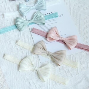 Puede incluir: Varios lazos de colores pastel están dispuestos sobre una superficie blanca. Los lazos son en tonos de azul claro, verde menta, rosa y crema. Las cintas para el pelo tienen bandas elásticas. Los lazos tienen un aspecto brillante y texturizado.