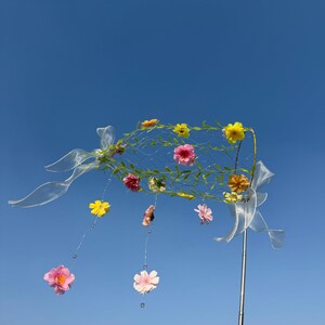May include: A decorative floral arrangement against a clear blue sky. The arrangement features a wire frame adorned with colorful flowers, green vines, and clear ribbons. Several flowers hang from the frame with clear strings and beads.