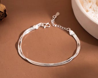 May include: A silver bracelet with three delicate strands. The bracelet has a simple clasp and adjustable chain. The jewelry is displayed on a brown surface with dried flowers and a white bowl in the background.