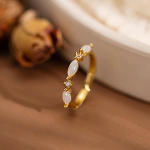 Peut inclure: Une bague dorée ornée d'une série de pierres précieuses blanches de forme ovale et de petits accents transparents. La bague est posée sur un fond doux et neutre, soulignant le design délicat du bijou. L'anneau de la bague est fin et poli.