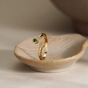 May include: A gold open-ended ring featuring two circular settings at the ends. One setting contains a small, round, vibrant green gemstone, while the other setting holds a small, round, clear white gemstone. The band is polished gold.