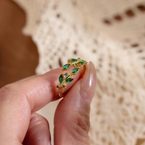 Op de afbeelding: Een goudkleurige ring met een bladerenontwerp, versierd met groene edelstenen. De ring wikkelt zich om de vinger en heeft een delicate rangschikking van bladeren en smaragdgroene stenen. De ring wordt door een hand omhoog gehouden.