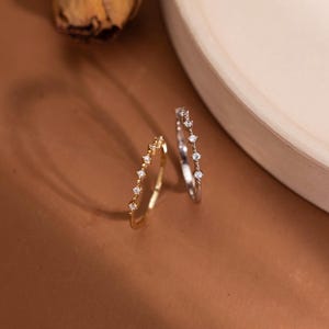 May include: Two thin rings, one in gold tone and one in silver tone, each featuring a series of small, clear, round-cut stones set along the band. The rings are positioned upright, showcasing their delicate metal construction and sparkling stones.