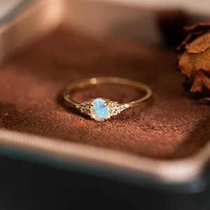 May include: A gold ring featuring a central oval-shaped iridescent opal stone flanked by two small clear accent stones on each side. The thin, polished gold band holds the stones in a prong setting, creating a delicate and refined appearance.