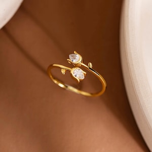 May include: A gold ring with a floral design. The ring features two clear, teardrop-shaped gemstones. The ring is set against a brown background with a white curved edge in the upper right corner.