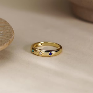 May include: A gold band ring featuring three star-shaped settings, each containing a small gemstone. The stones are coloured green, clear, and blue, and are arranged horizontally along the centre of the polished, smooth, rounded metal band.