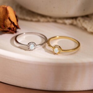 Pode incluir: Dois anéis finos, um em metal prateado e outro em metal dourado, cada um com uma pequena pedra central redonda de opala iridescente rodeada por um halo de minúsculas pedras de destaque claras e brilhantes numa banda polida e lisa.