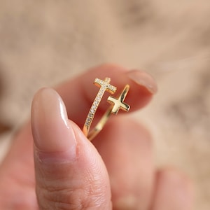Peut inclure: Une bague dorée ornée de deux croix. L'une est ornée de petites pierres scintillantes, l'autre est une croix simple. La bague est tenue entre les doigts, soulignant son design délicat. Idéale pour un usage religieux.