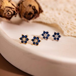 Op de afbeelding: Twee paar bloemvormige oorknopjes met zes donkerblauwe bloemblaadjes rondom een enkele heldere steen in het midden. De oorbellen zijn voorzien van een goudkleurige metalen zetting, wat zorgt voor een heldere en glanzende uitstraling.