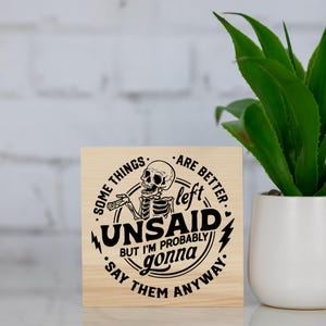 May include: A wooden sign featuring a black and white skeleton design and the phrase "Some things are better left unsaid but I'm probably gonna say them anyway." A green plant in a white pot is to the right.