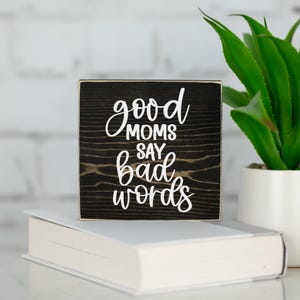 May include: A small, square wooden sign with the phrase "good moms say bad words" in white script. The sign is black with visible wood grain and sits atop a white book. A potted green plant is in the background.