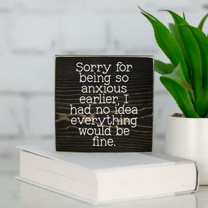May include: A black wooden block with white text that reads "Sorry for being so anxious earlier, I had no idea everything would be fine." The block is on a white book, with a green plant in a white pot in the background.