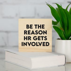 May include: A wooden block with the text "BE THE REASON HR GETS INVOLVED" in black lettering. The block is sitting on a white book. A green plant in a white pot is in the background.