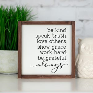 Op de afbeelding: Een houten bord met een witte achtergrond en een donkerbruine lijst. Het bord toont de woorden "be kind, speak truth, love others, show grace, work hard, be grateful, always" in zwart schrift. Een decoratieve uil en een potplant staan op de achtergrond.