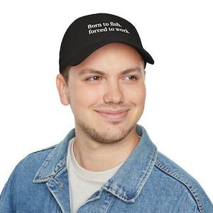 Può includere: Cappellino da baseball nero con la scritta bianca "Born to fish, forced to work". Il cappellino è indossato da una persona con una giacca di jeans.