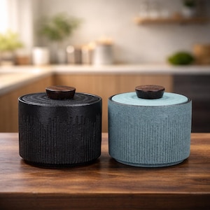 May include: Two ceramic canisters with wooden lids. One is black, the other is a muted teal. Both have a textured, vertically-lined design. The canisters sit on a wooden surface, suggesting kitchen or home decor.