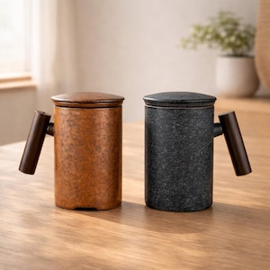 Puede incluir: Dos tazas de cerámica con tapas y asas de madera. Una taza es negra moteada, la otra marrón moteada. Ambas tazas son cilíndricas y están sobre una superficie de madera.