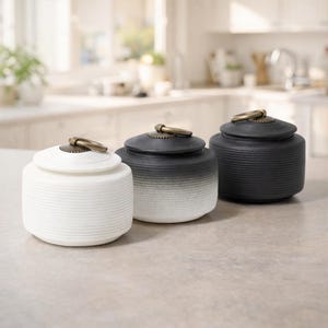 May include: Three ceramic jars with lids. The jars are white, black and white gradient, and black. Each jar has a ribbed texture and a brass-coloured ring handle on the lid. The jars are arranged on a light-coloured countertop.