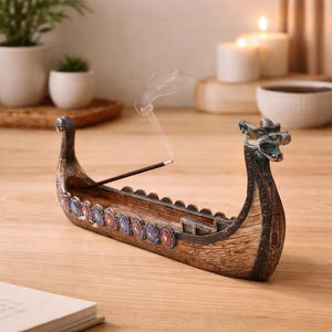 Puede incluir: Un porta incienso de madera con forma de barco vikingo, con una cabeza de dragón. El porta incienso es marrón con detalles azules, rojos y dorados. El humo del incienso se eleva. El porta incienso está sobre una superficie de madera.