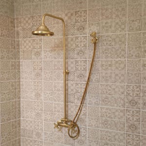 May include: A wall-mounted shower system in a brushed brass finish, featuring a fixed overhead shower head, a flexible handheld shower wand with a hose, and two cross-handle valves for water control, all mounted on a tiled wall surface.