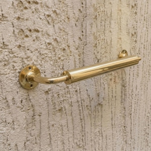 May include: A polished brass towel rail fixed to a textured, off-white wall. The rail has a cylindrical design with decorative detailing near the end and a circular mounting plate. The fixture is designed for bathroom use.
