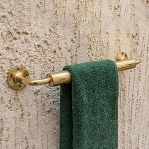 May include: A brass towel rail with a green towel hanging from it. The rail is mounted on a textured wall. The towel rail has a circular base and a curved arm.