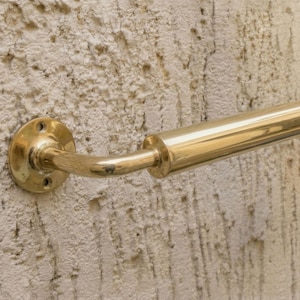 May include: A polished brass handrail fixed to a textured wall. The handrail features a curved section and a cylindrical bar. The wall has a rough, off-white surface, typical of a rendered finish.