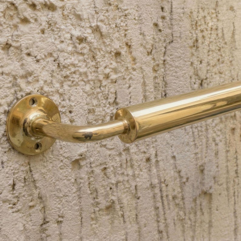 May include: A polished brass handrail fixed to a textured wall. The handrail features a curved section and a cylindrical bar. The wall has a rough, off-white surface, typical of a rendered finish.