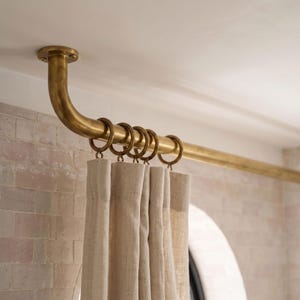 May include: A ceiling-mounted brass curtain rod with a curved end, featuring a series of circular rings attached to the rod. A light-colored, textured fabric curtain hangs from the rings, creating vertical folds along the length of the material.
