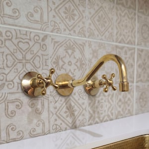 May include: A wall-mounted brass faucet featuring a curved spout and two cross-handle controls. The fixture has a polished metallic finish and is installed against a wall with light-colored square tiles that display a repeating geometric pattern.