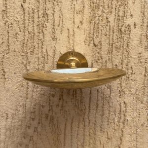 May include: A brass soap dish with a circular design, mounted on a textured wall. The dish has a shallow, curved shape with a white interior and a brass backplate. The fixture is secured with a single screw.