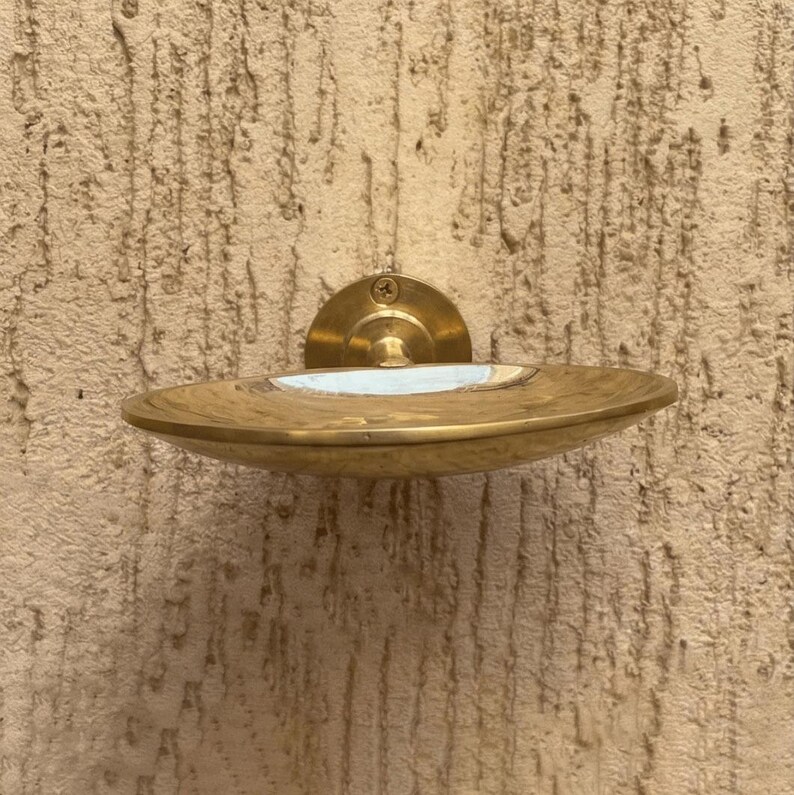 May include: A brass soap dish with a circular design, mounted on a textured wall. The dish has a shallow, curved shape with a white interior and a brass backplate. The fixture is secured with a single screw.