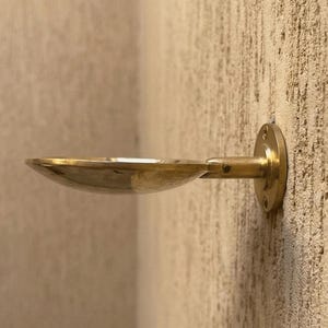 May include: A brass wall-mounted soap dish with a shallow, oval bowl and a short, horizontal arm. The dish is attached to the wall with a circular mounting plate. The finish is a warm, metallic gold.