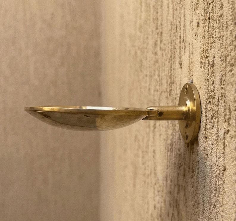 May include: A brass wall-mounted soap dish with a shallow, oval bowl and a short, horizontal arm. The dish is attached to the wall with a circular mounting plate. The finish is a warm, metallic gold.
