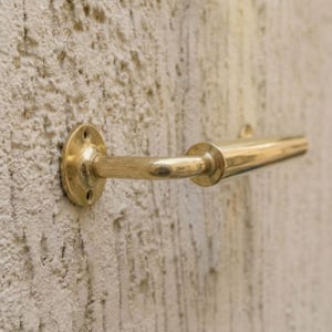 May include: A brass-coloured towel bar fixed to a wall. The bar features a circular mounting plate with screw holes, a curved arm, and a cylindrical bar. The finish is shiny and reflects light. The bar is mounted on a textured, light-coloured wall.