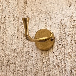 May include: A brass wall hook with a curved arm and a circular base. The hook is mounted on a textured, off-white wall. The hook's design features a flared top and a rounded end, suitable for hanging items.