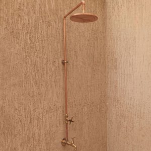 Pode incluir: Um sistema de duche de cobre montado na parede com um tubo vertical, um chuveiro circular, uma válvula de controlo com manípulo em cruz e uma torneira inferior. O acessório metálico tem um acabamento quente em tons de castanho-avermelhado.