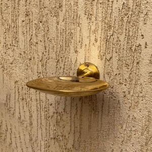 May include: A brass-colored, wall-mounted soap dish. The dish has a shallow, oval shape with a raised rim and a small, circular indentation in the center. The dish is attached to the wall with a round, brass-colored mounting plate.