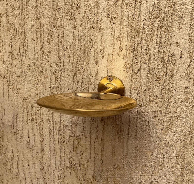 May include: A brass-colored, wall-mounted soap dish. The dish has a shallow, oval shape with a raised rim and a small, circular indentation in the center. The dish is attached to the wall with a round, brass-colored mounting plate.