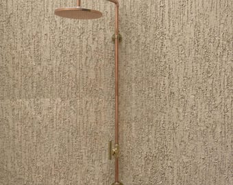 May include: A wall-mounted shower system featuring a circular shower head and a vertical pipe made of copper. The fixture includes a brass-coloured handle for water control and is secured to the wall with two circular mounting plates at the top and bottom.