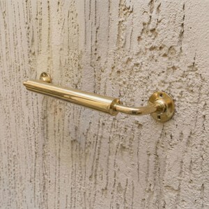 May include: A polished brass door handle, featuring a curved lever and a round mounting plate. The handle is mounted on a textured, off-white wall. The design includes a cylindrical grip and decorative detailing.