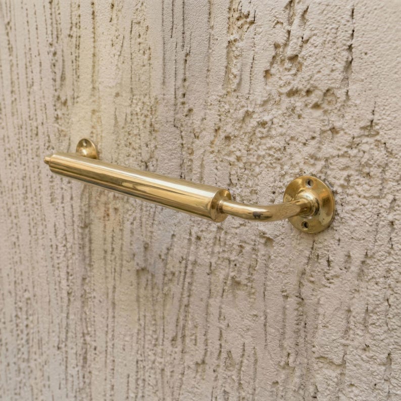 May include: A polished brass door handle, featuring a curved lever and a round mounting plate. The handle is mounted on a textured, off-white wall. The design includes a cylindrical grip and decorative detailing.