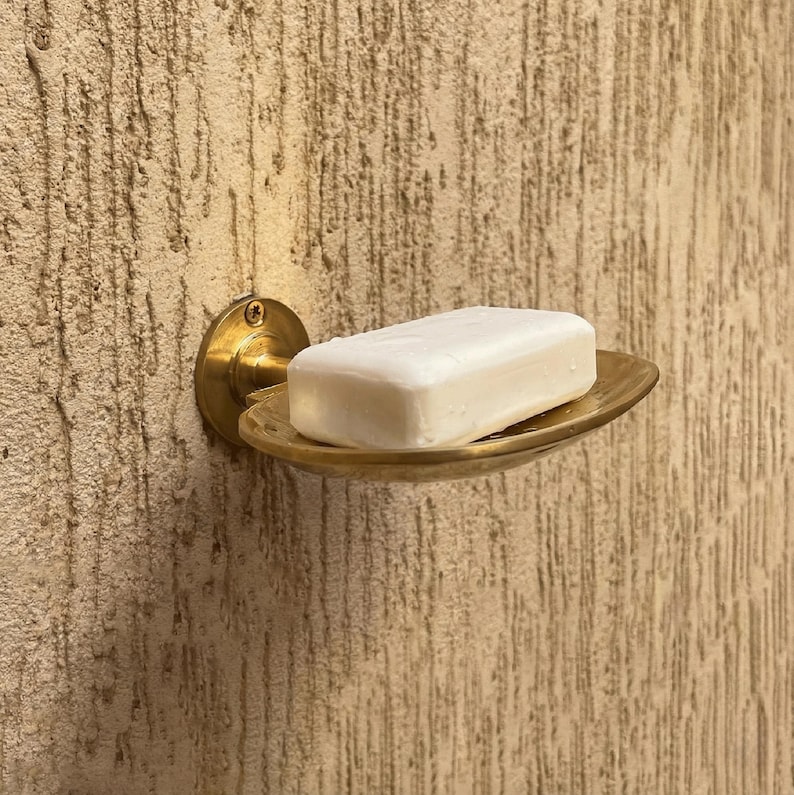 May include: A brass soap dish mounted on a textured wall. The dish holds a white bar of soap. The dish has a circular backplate and a shallow, oval-shaped holder. The wall has a rough, stucco-like finish.