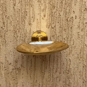 May include: A brass-toned soap dish with a circular backplate and a curved, oval-shaped dish. The dish has a light-colored interior and is mounted on a textured wall. The fixture is secured with a single screw.