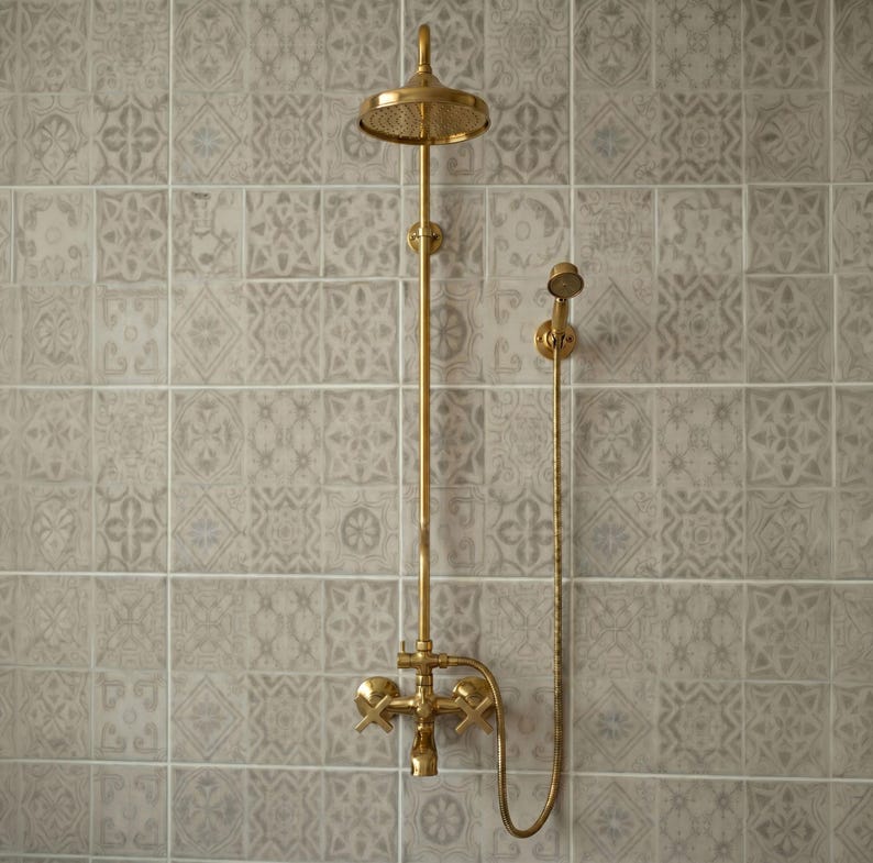 May include: A wall-mounted brass shower system featuring a circular overhead shower head, a vertical pipe, a handheld shower attachment with a flexible hose, and two cross-handle valves for controlling water flow and temperature.