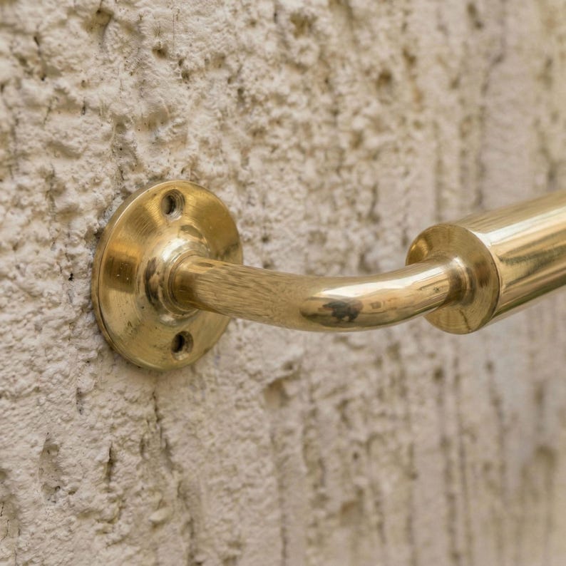May include: A brass handrail fixed to a textured, off-white wall. The handrail has a curved design, a circular mounting plate with three screw holes, and a cylindrical grip. The wall has a rough, uneven surface.