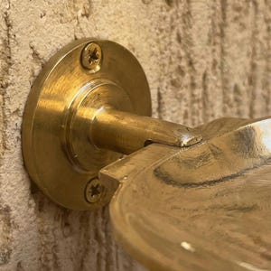 May include: Close-up of a brass wall-mounted soap dish. The dish has a circular base with three screws and a curved arm supporting a shallow, rounded dish. The brass has a polished finish, and the dish is mounted on a textured wall.