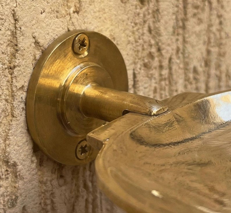 May include: Close-up of a brass wall-mounted soap dish. The dish has a circular base with three screws and a curved arm supporting a shallow, rounded dish. The brass has a polished finish, and the dish is mounted on a textured wall.